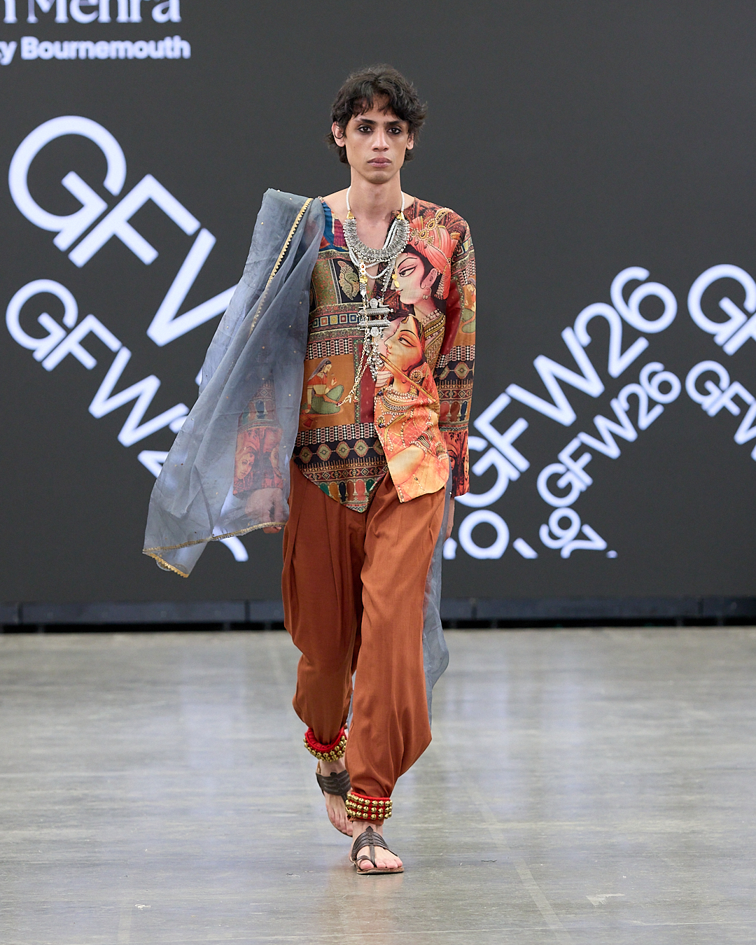A model walks the runway wearing a colourful patterned top, loose brown trousers and a sheer blue drape, with a dark backdrop behind them.