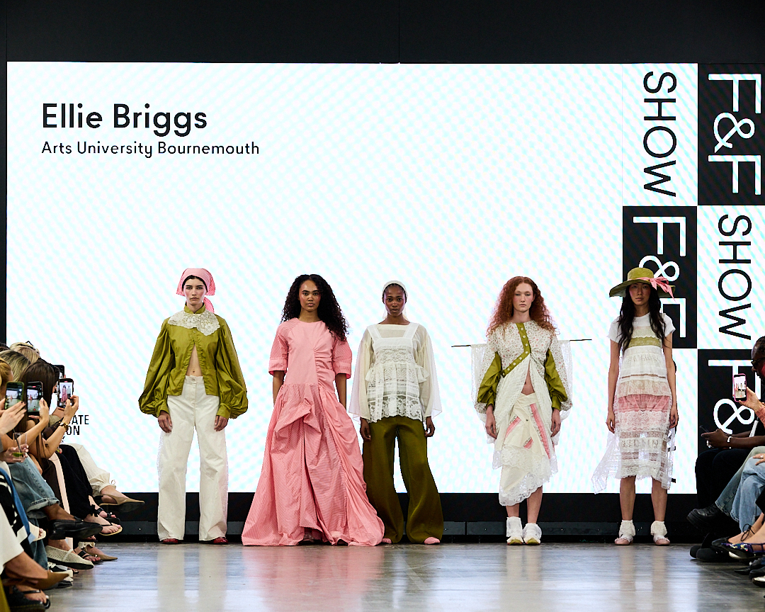Five models walk a runway in assorted pastel and neutral outfits at a fashion show, with 'Ellie Briggs' and 'Arts University Bournemouth' displayed on the screen behind them.