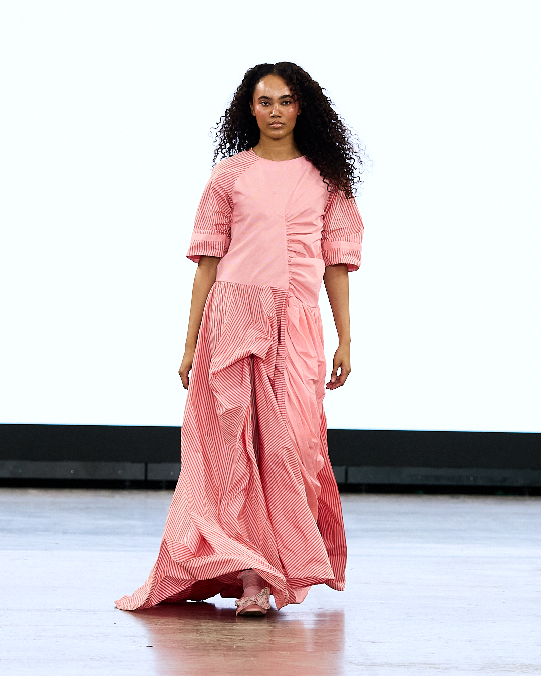 A woman with curly hair wears a pink patterned dress on a runway with a plain white backdrop.