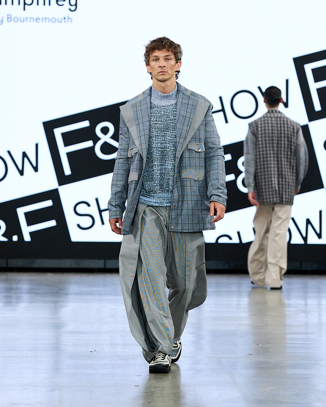 A male model wearing a layered grey outfit walks down a fashion runway, with another model in a checked jacket in the background.