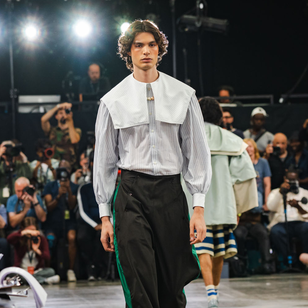 Male model on runway wearing white shirt with large collar and black trousers with green stripe, audience and bright lights visible.