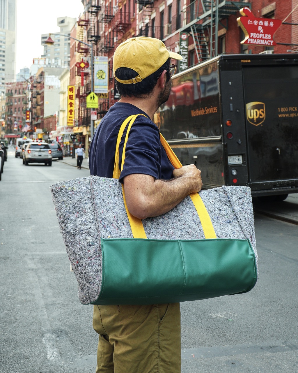 A man in a yellow cap carries a large grey and green tote bag across a city street, with a UPS truck and shop signs in the background.