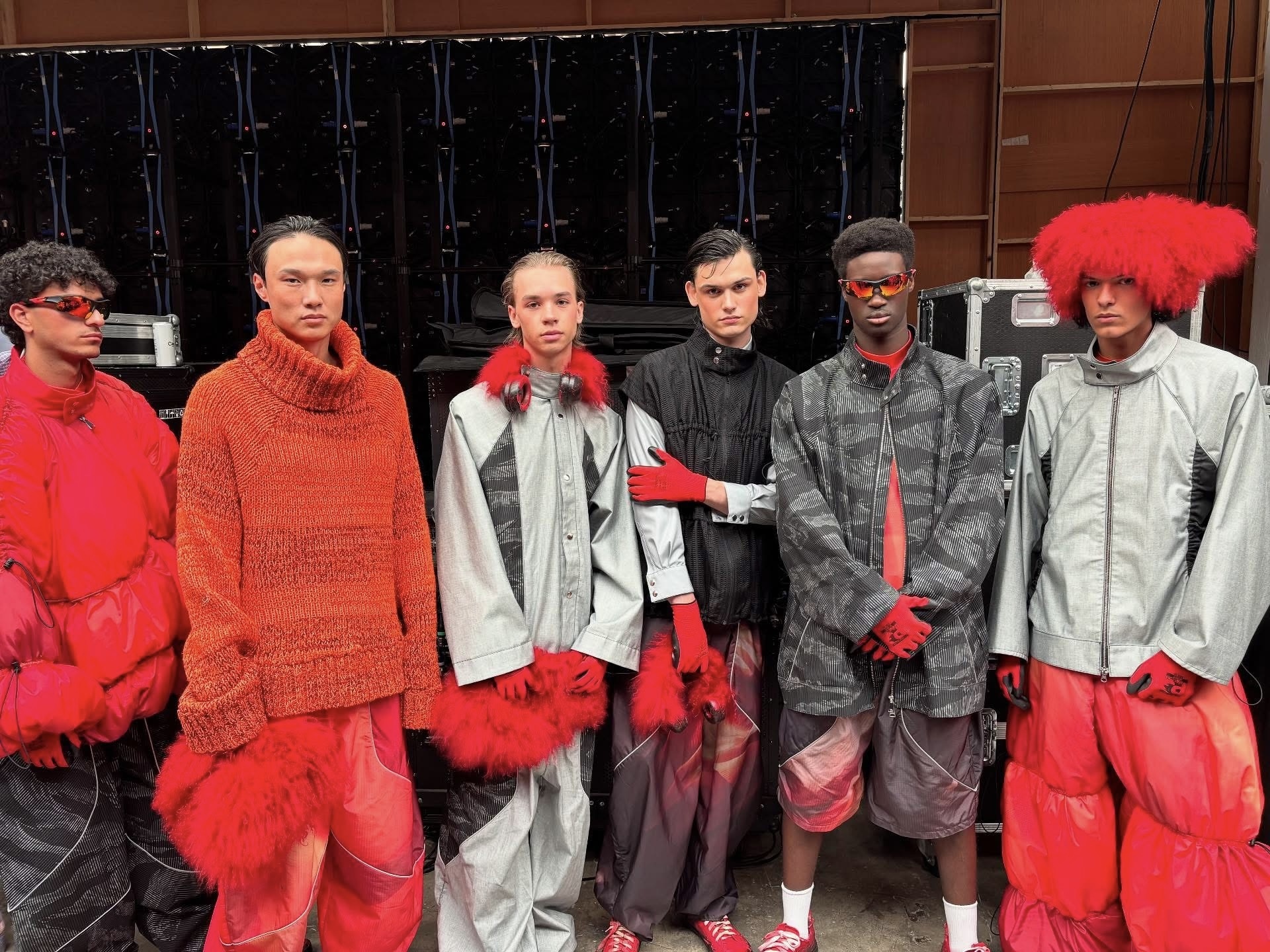 Six models in avant-garde fashion line up outdoors wearing oversized grey and red garments with dramatic red fur accessories.