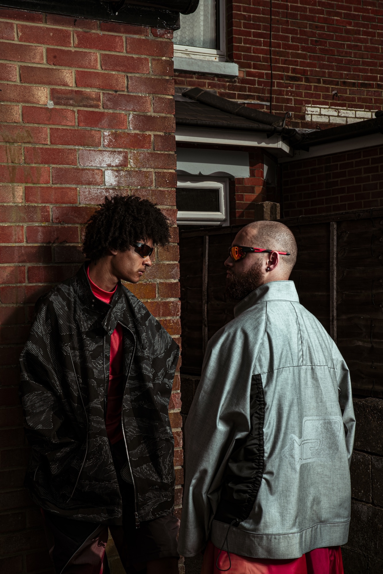 Two models in conversation outside red brick building. One wears dark patterned jacket with red accents, other in light blue shirt.
