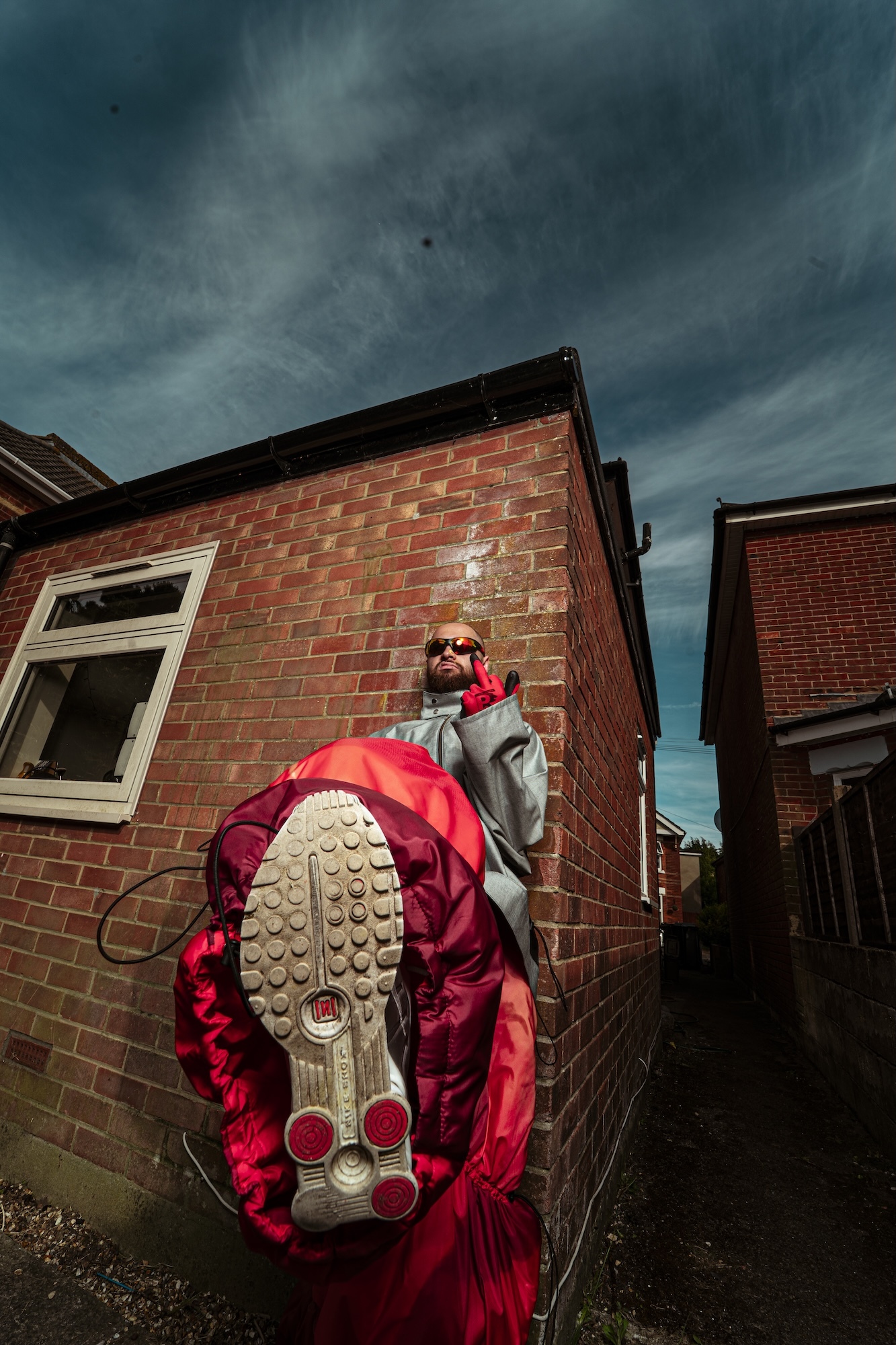 Person in red tracksuit and sunglasses jumping mid-air showing trainer soles, against red brick houses and stormy grey sky.