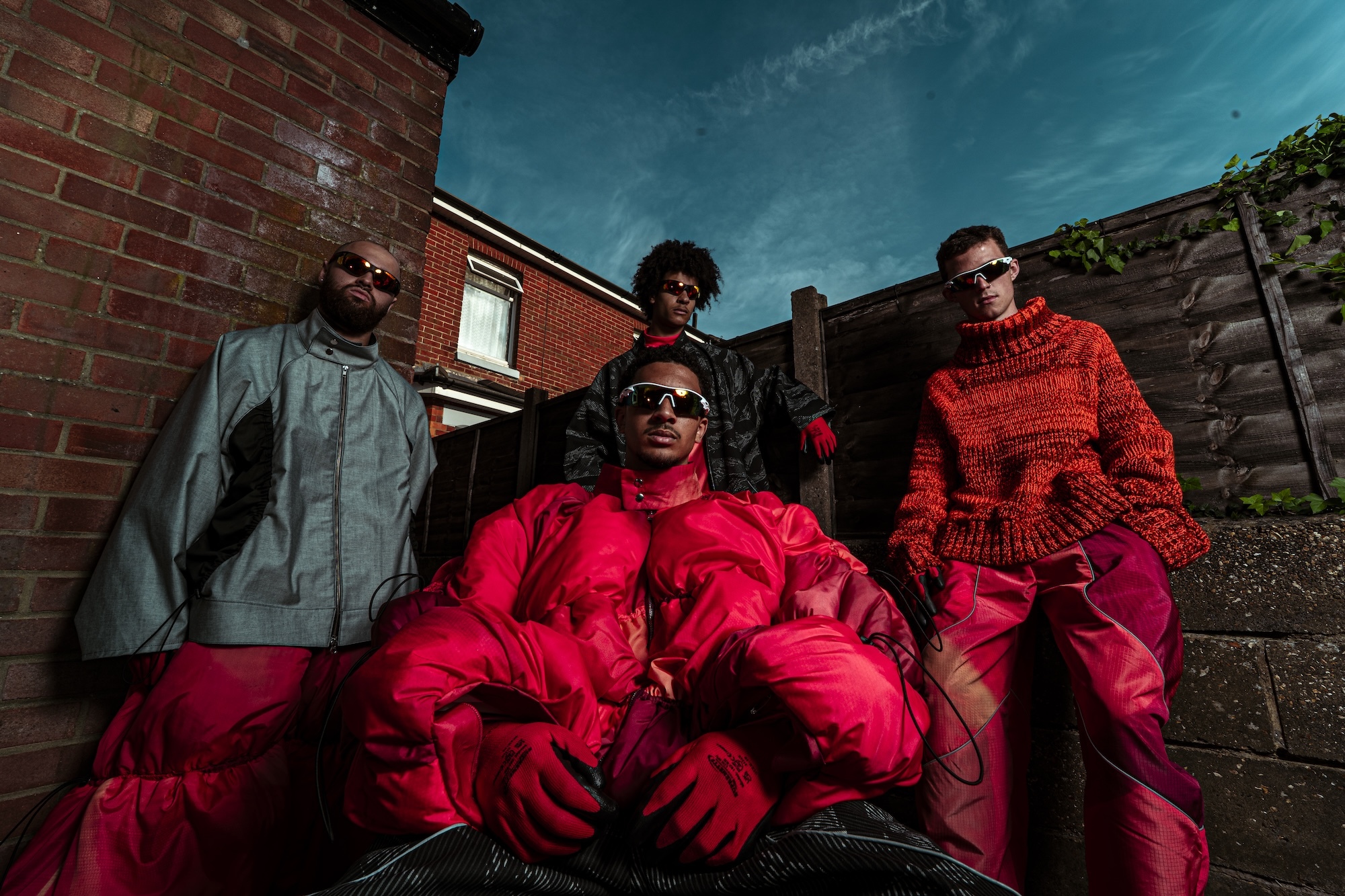 Four people in bright red tracksuits and sunglasses stand against brick buildings under turquoise sky with white clouds.