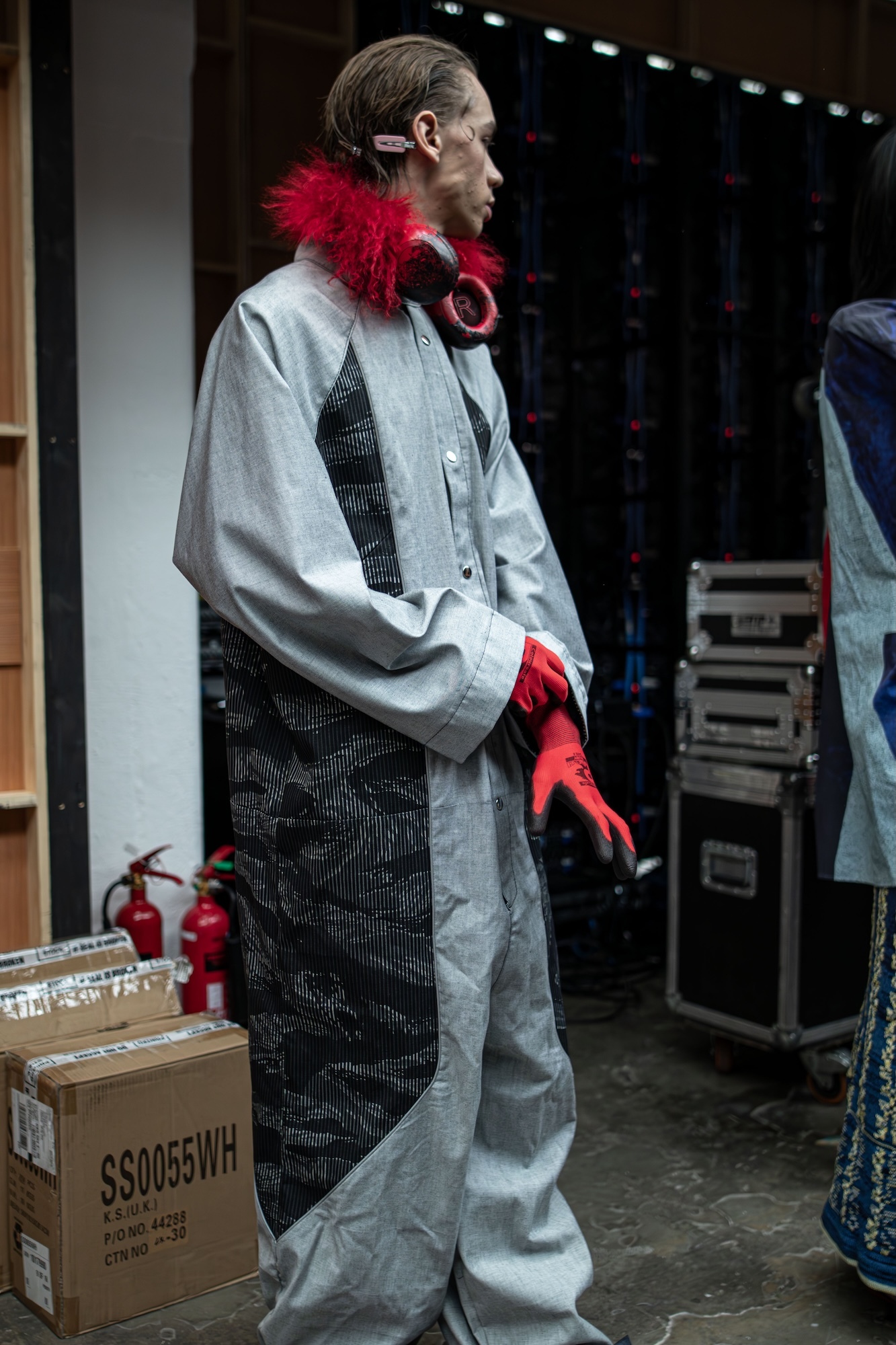 Man in grey coveralls with red fur collar and black patterned vest, wearing red headphones, standing near cardboard boxes backstage.