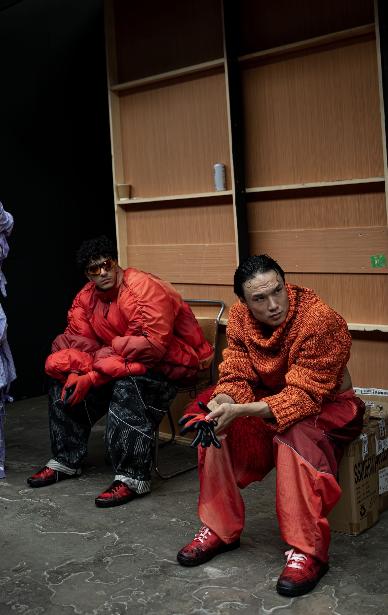 Two models in bright red costumes and knitted jumpers sit on wooden boxes backstage, with blonde person partially visible on left.