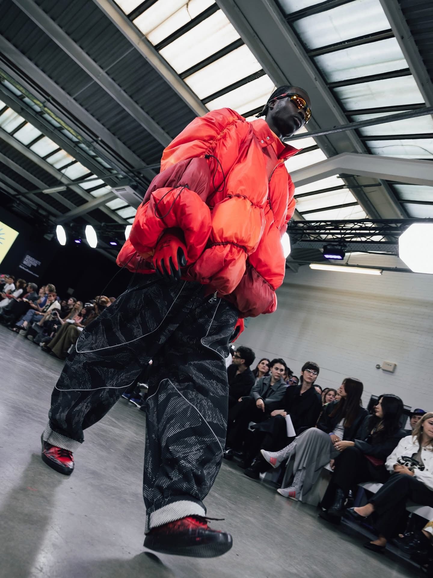 Model in oversized red puffer jacket and dark trousers walks runway at fashion show with seated audience and industrial ceiling.