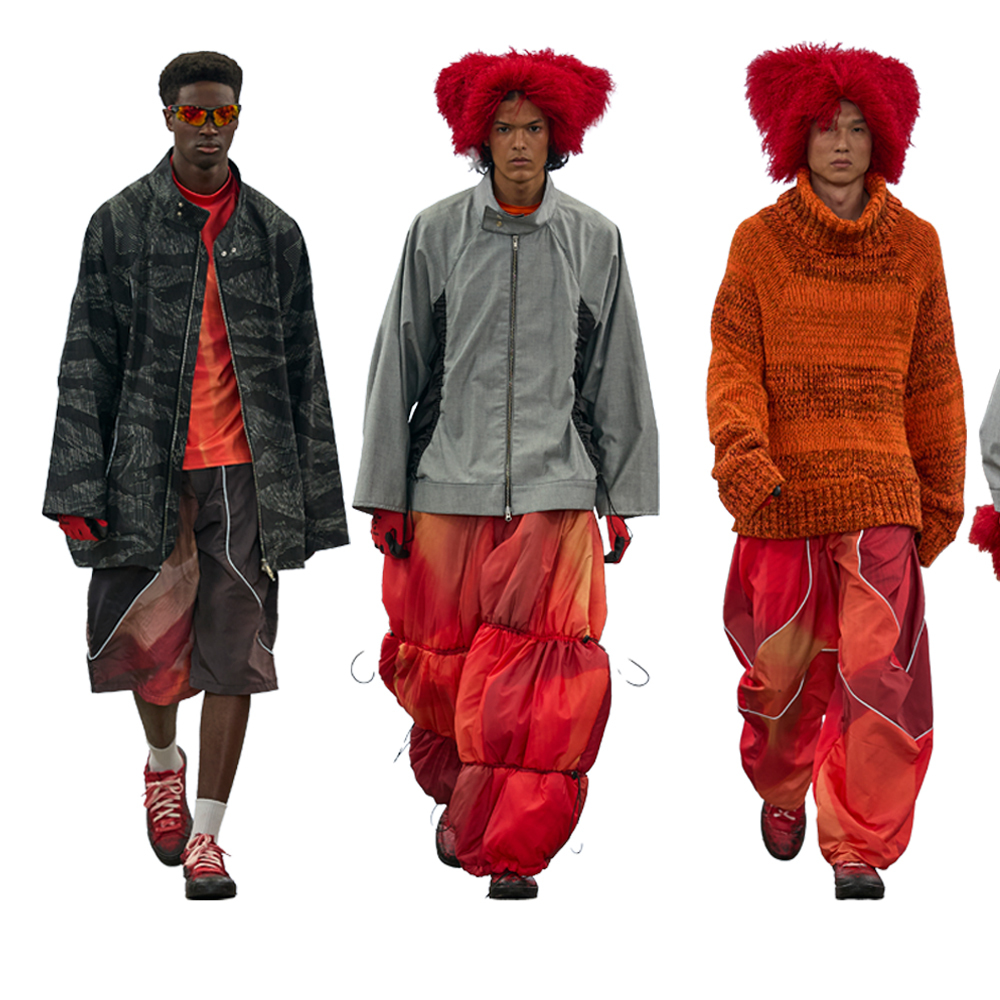 Three male models on runway wearing avant-garde menswear: dark coat with red accents, grey jacket with red fuzzy hat, orange knit jumper with red hat.