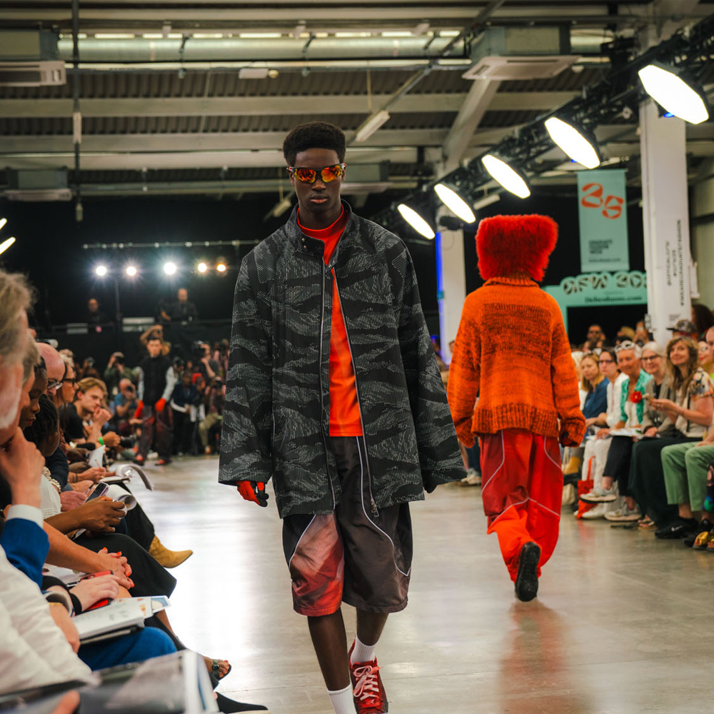 Models on runway wearing dark grey military-style jacket with red top and orange hooded jumper, audience seated on both sides.