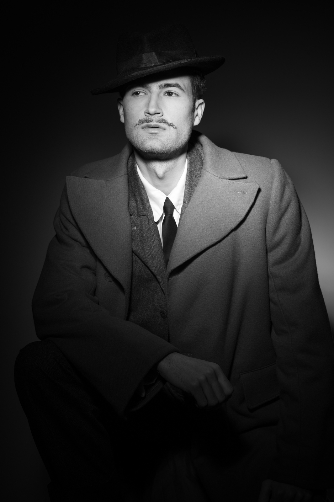 Black and white portrait of man wearing fedora hat, overcoat, tie and waistcoat, looking upward against dark background.