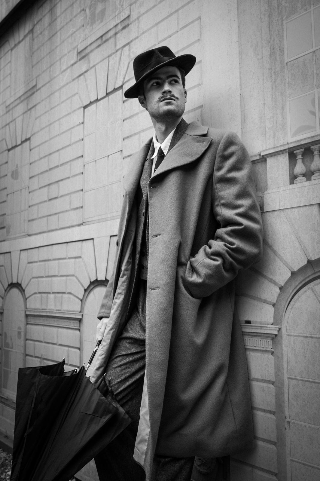 Man in black hat and long coat holding briefcase stands against stone building facade in black and white photograph.