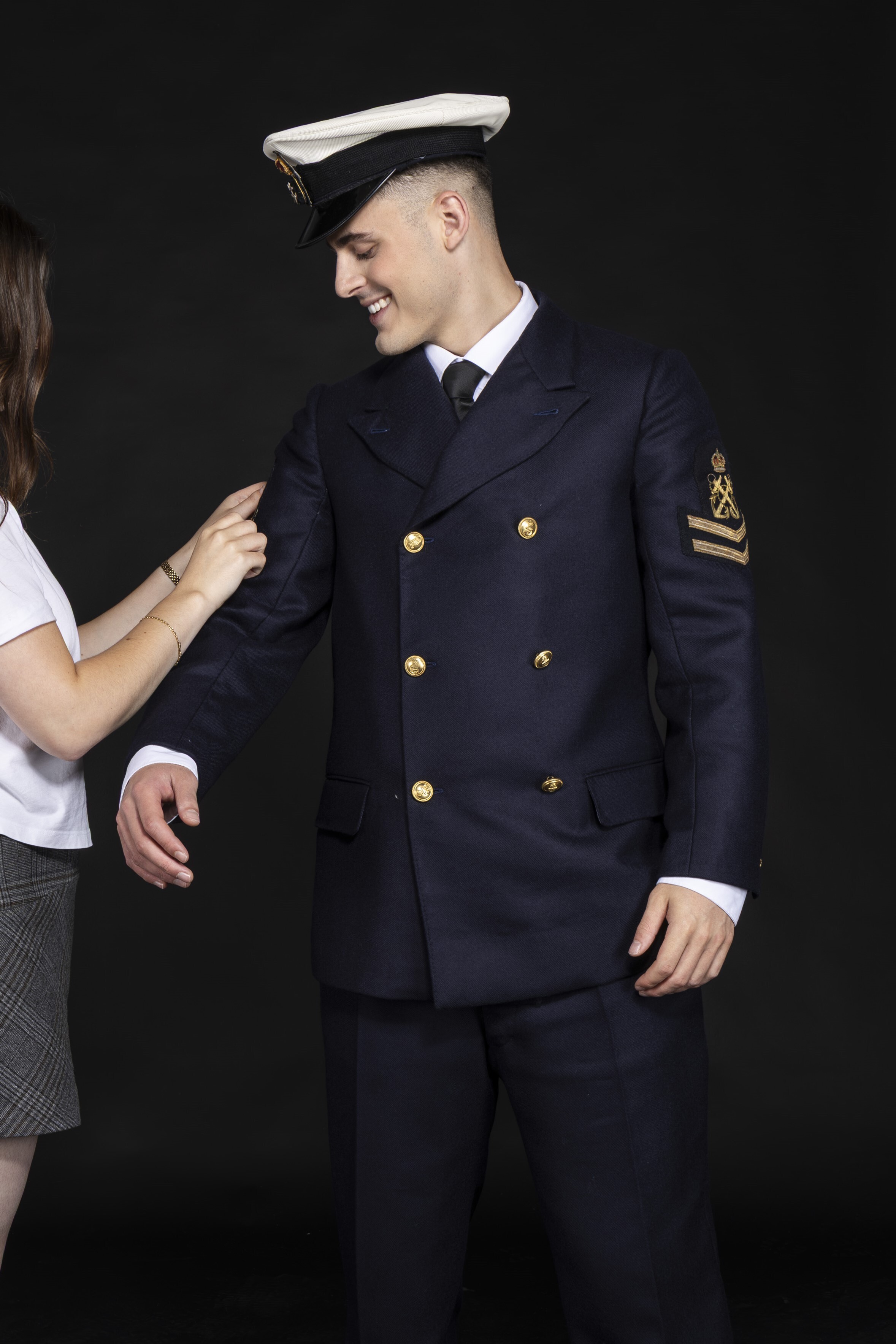 Young man in navy uniform with white peaked cap, gold buttons and insignia, smiling in profile against dark background.