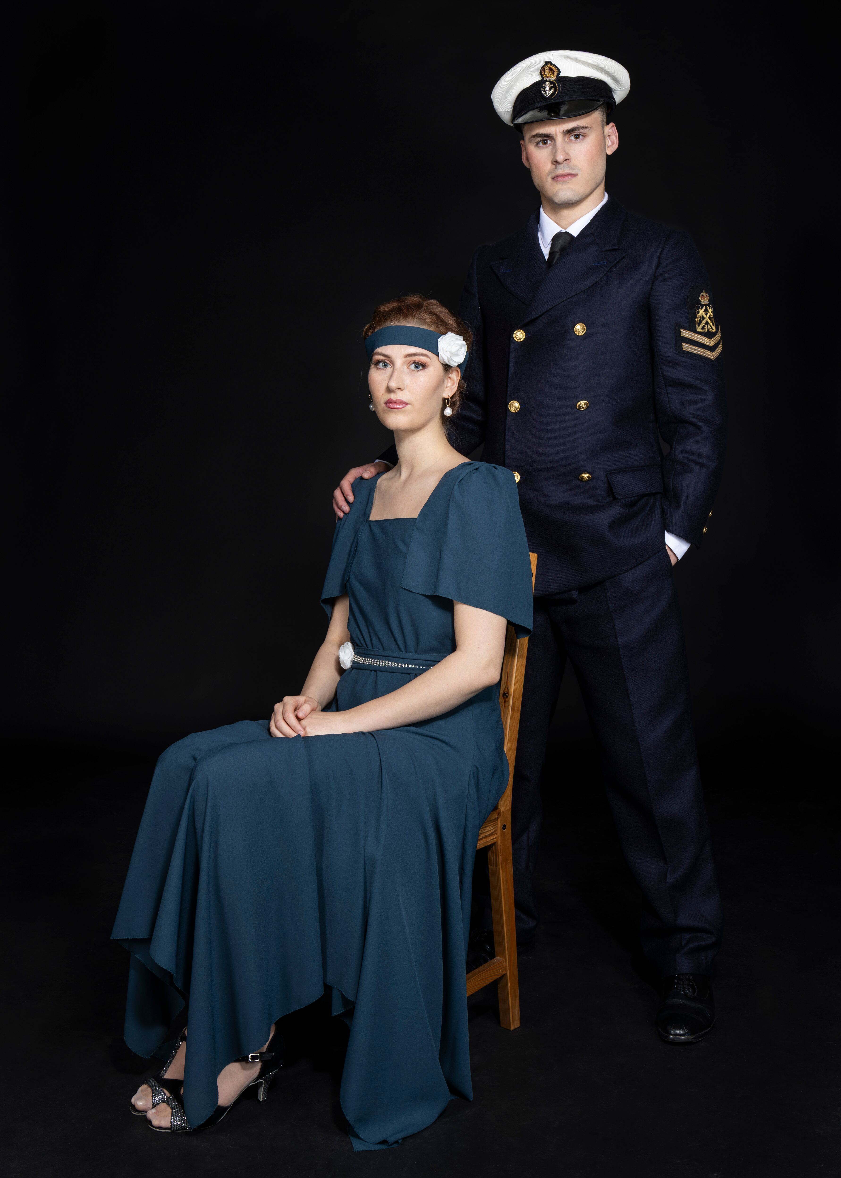 Woman in blue period dress seated on wooden chair with man in naval uniform standing behind her, both against black background.