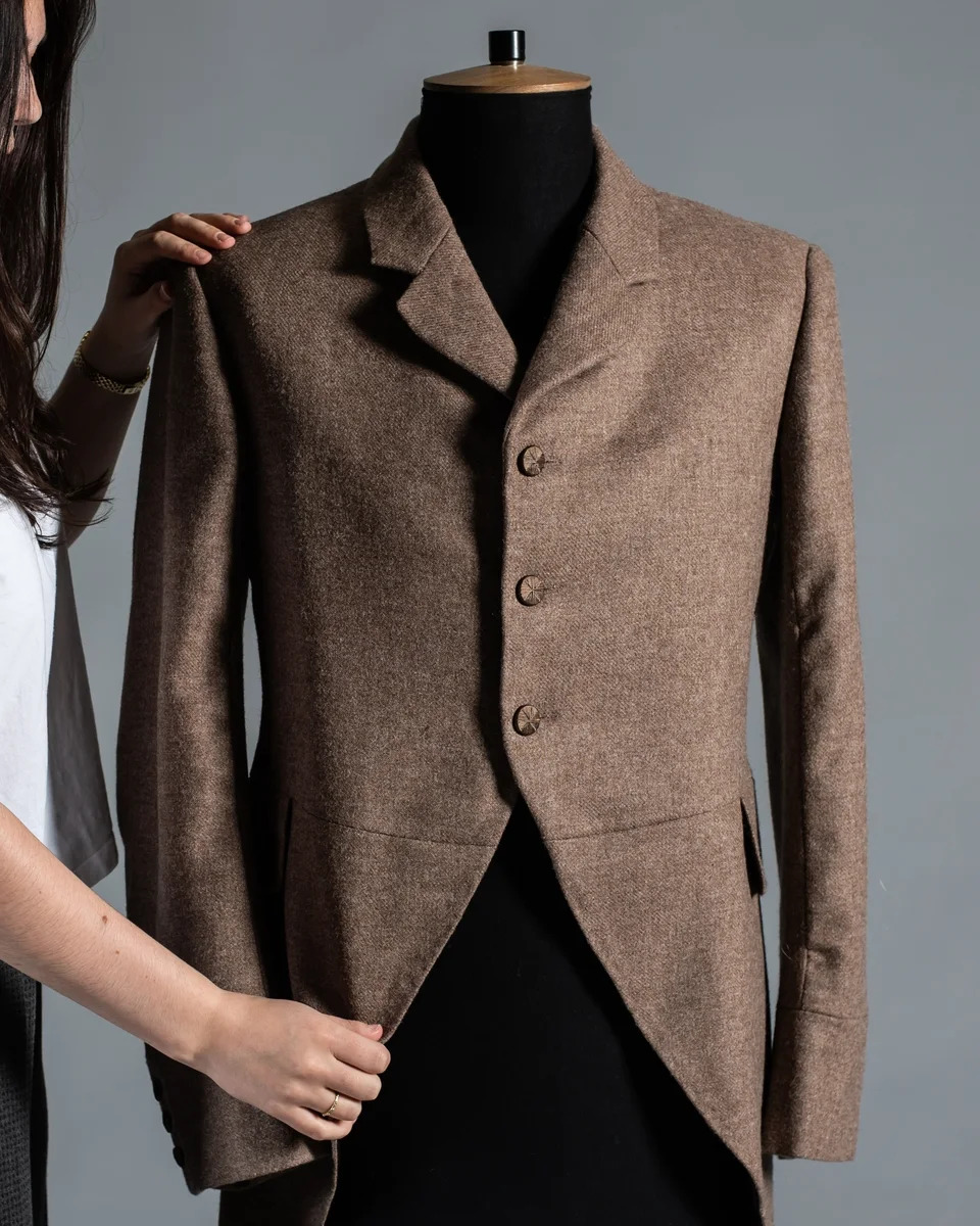 Brown tweed jacket with buttons on black mannequin, woman's hands adjusting the garment against grey background.