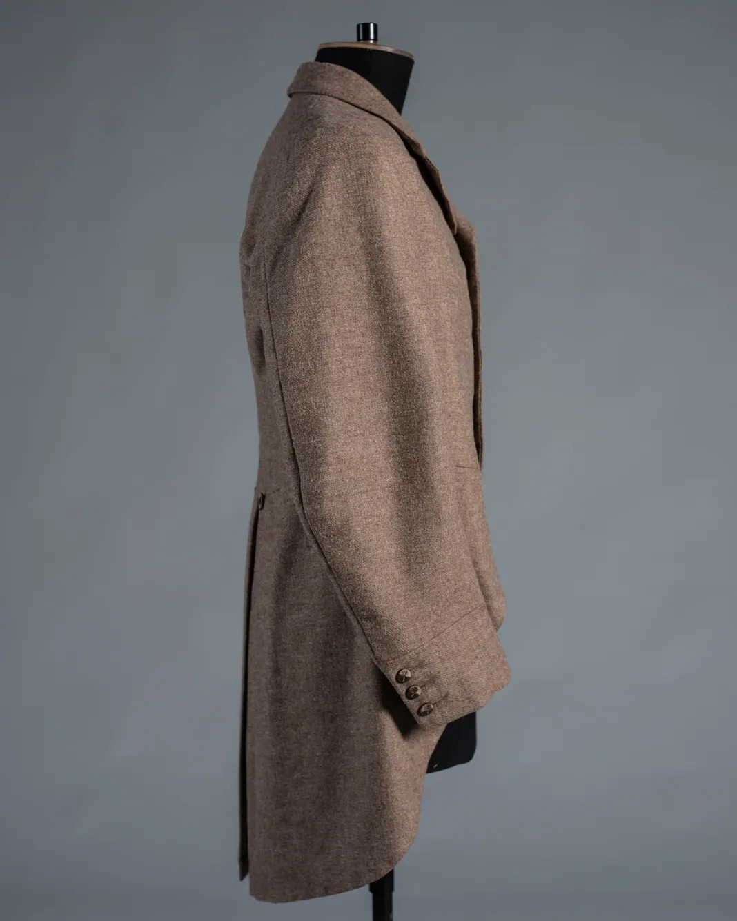 Side view of beige wool coat on black mannequin against grey background. Coat features lapels, buttons on sleeves, and mid-length cut.