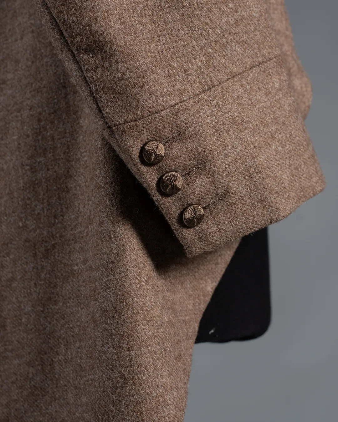 Close-up of brown tweed jacket sleeve with three decorative brown buttons on cuff, grey background visible.
