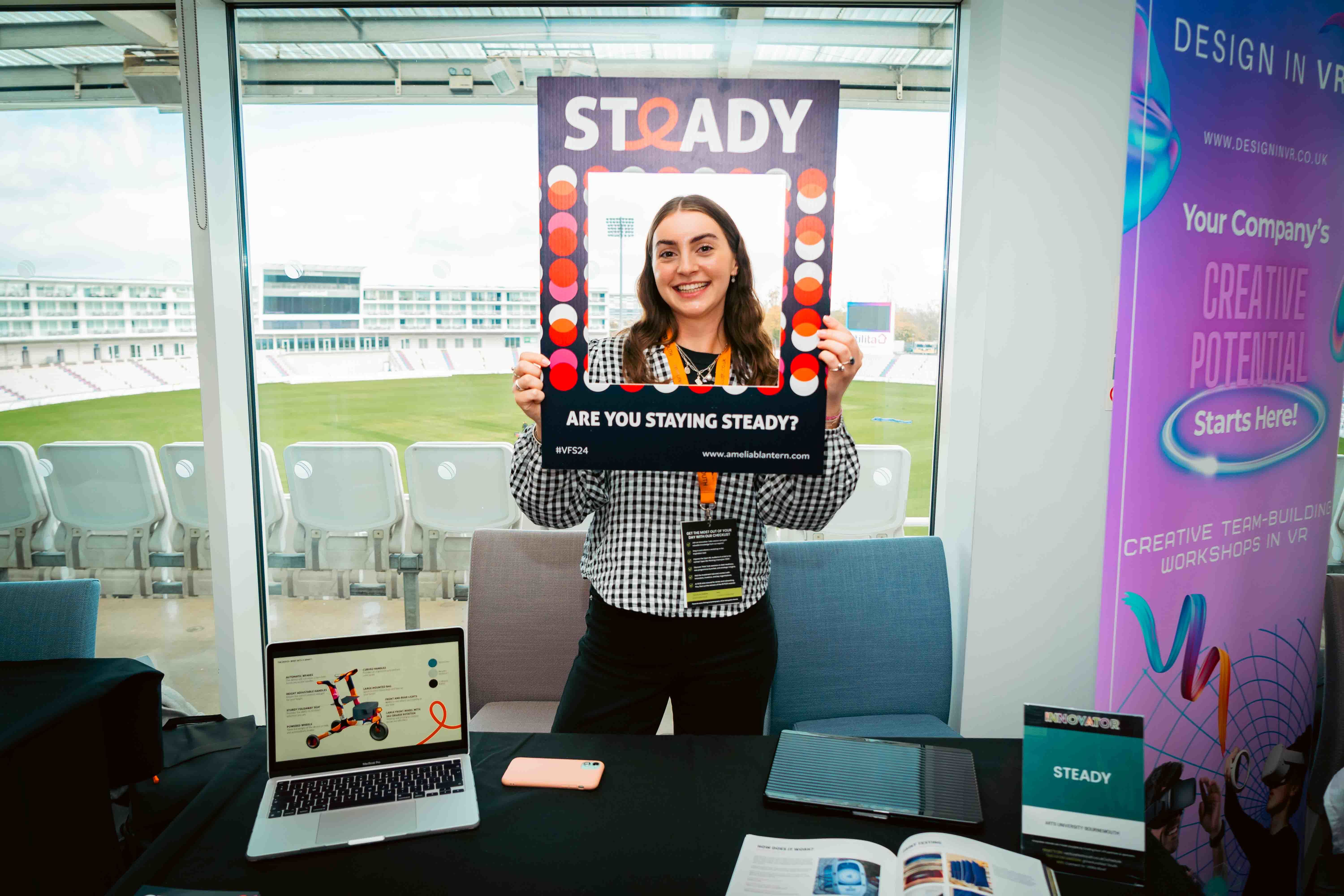 A. Blantern at her stall at Venturefest South 2024 holding a picture frame with the 'Steady' logo along the top and the slogan "Are you staying steady?" along the bottom.