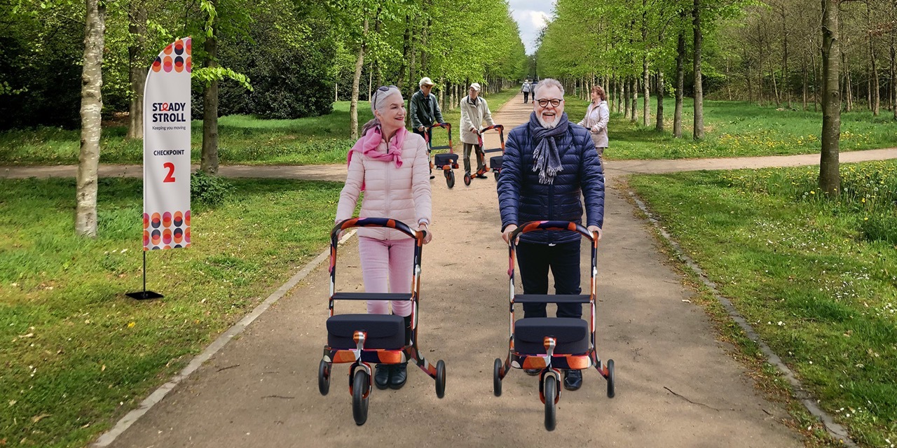 Two older adults smile while pushing colourful mobility aids along a park path.