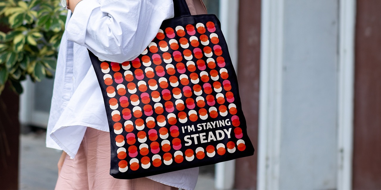 The side profile of a woman holding a colourful spotty tote bag over her shoulder which reads ‘I’m staying steady.’