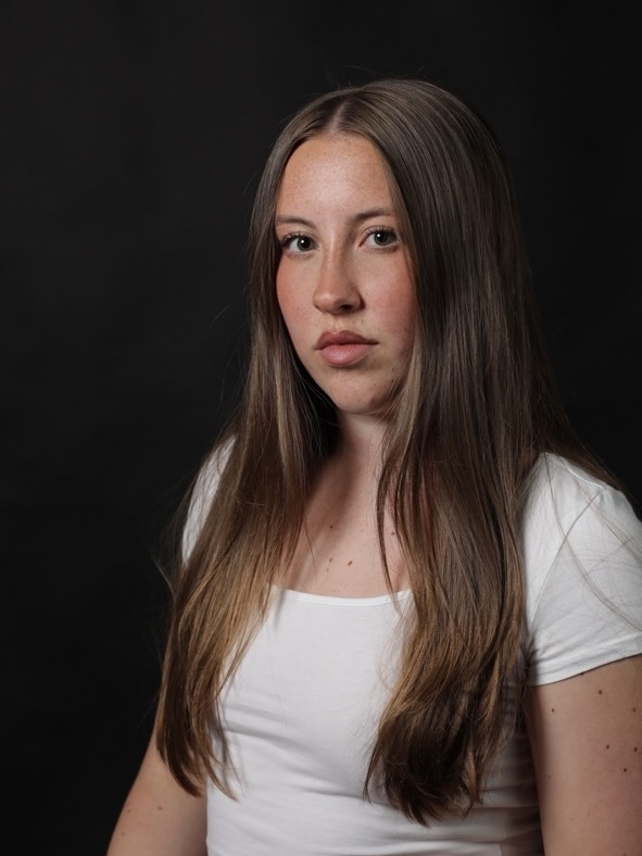 Woman with long brown hair wearing white t-shirt against black background, looking slightly to the side.