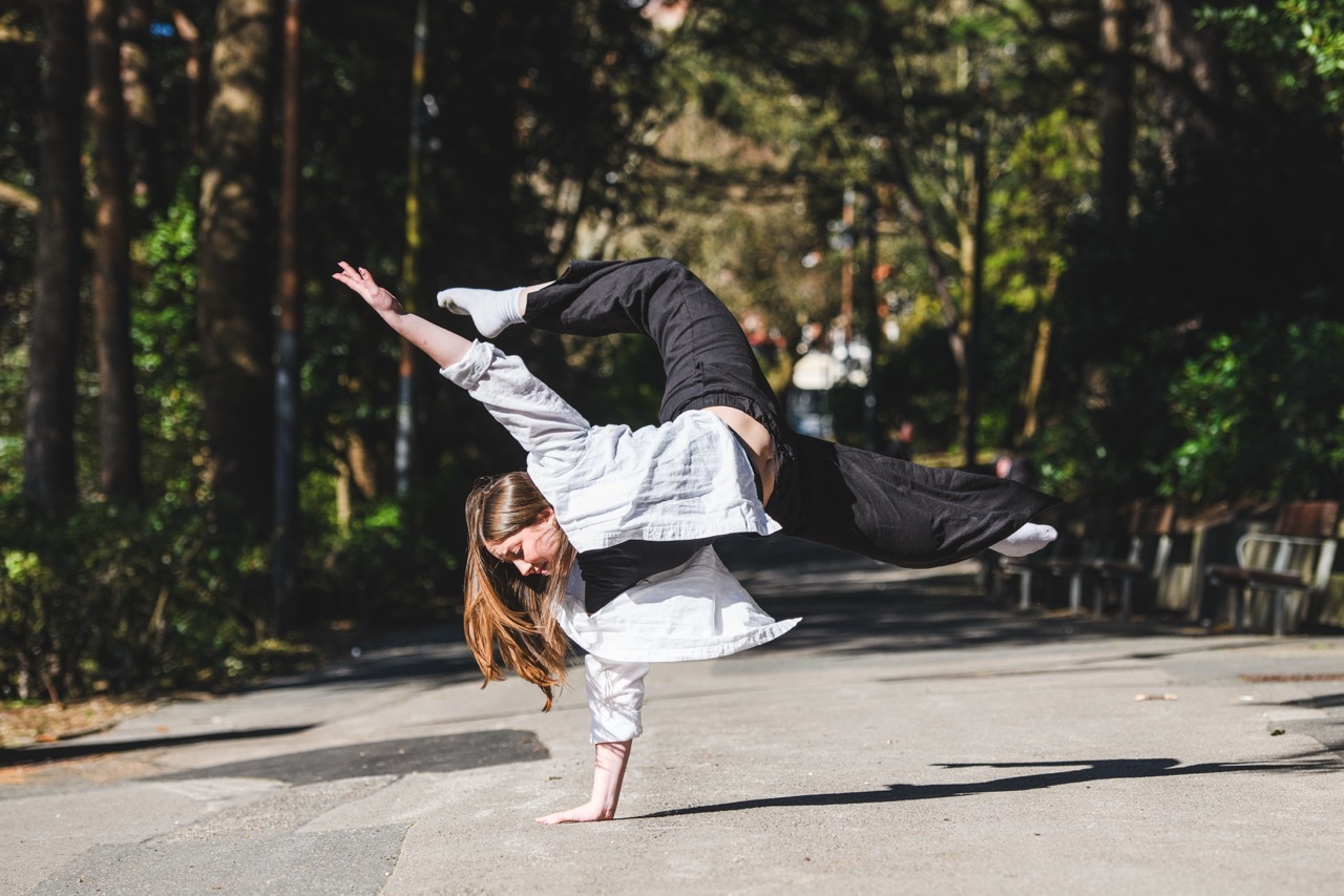 Woman in white top and black trousers performing breakdancing freeze move on one hand in park setting with trees and bench.