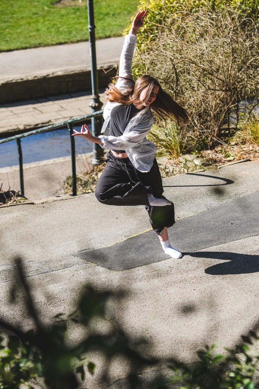 Woman with brown hair jumping mid-air on pavement, wearing grey hoodie and black trousers, with metal railings and bushes behind.