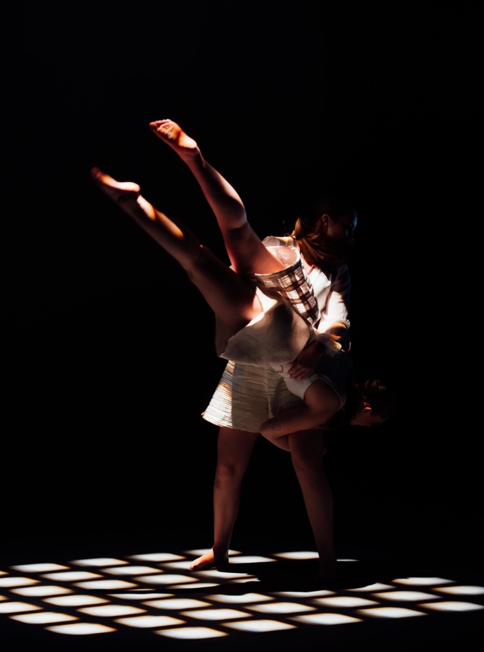 Two performers in dramatic lighting on black and white chequered floor, one lifting the other with arms raised against dark background.