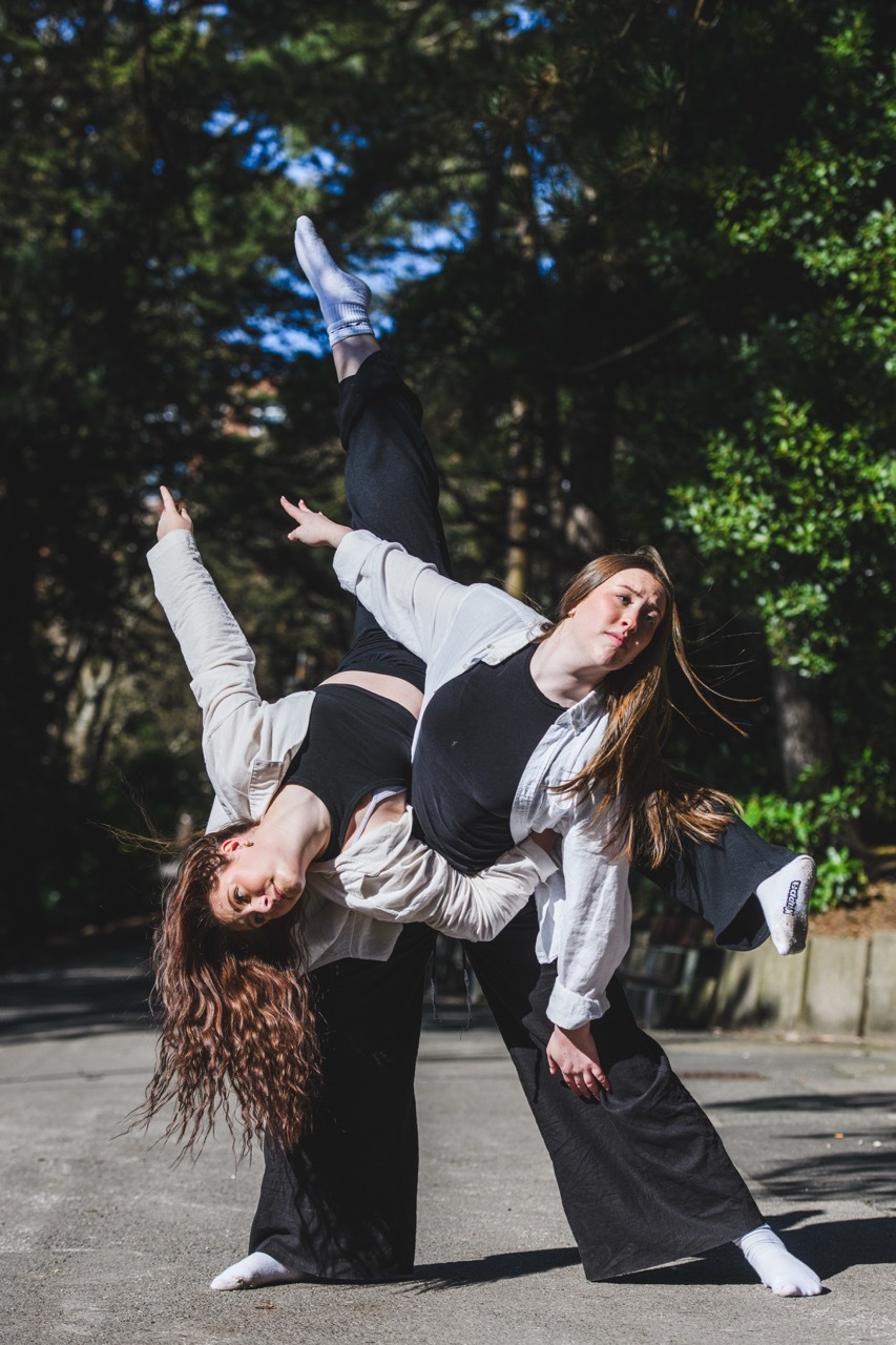 Two women performing contemporary dance outdoors, one supporting the other who is balanced horizontally with leg extended upward.