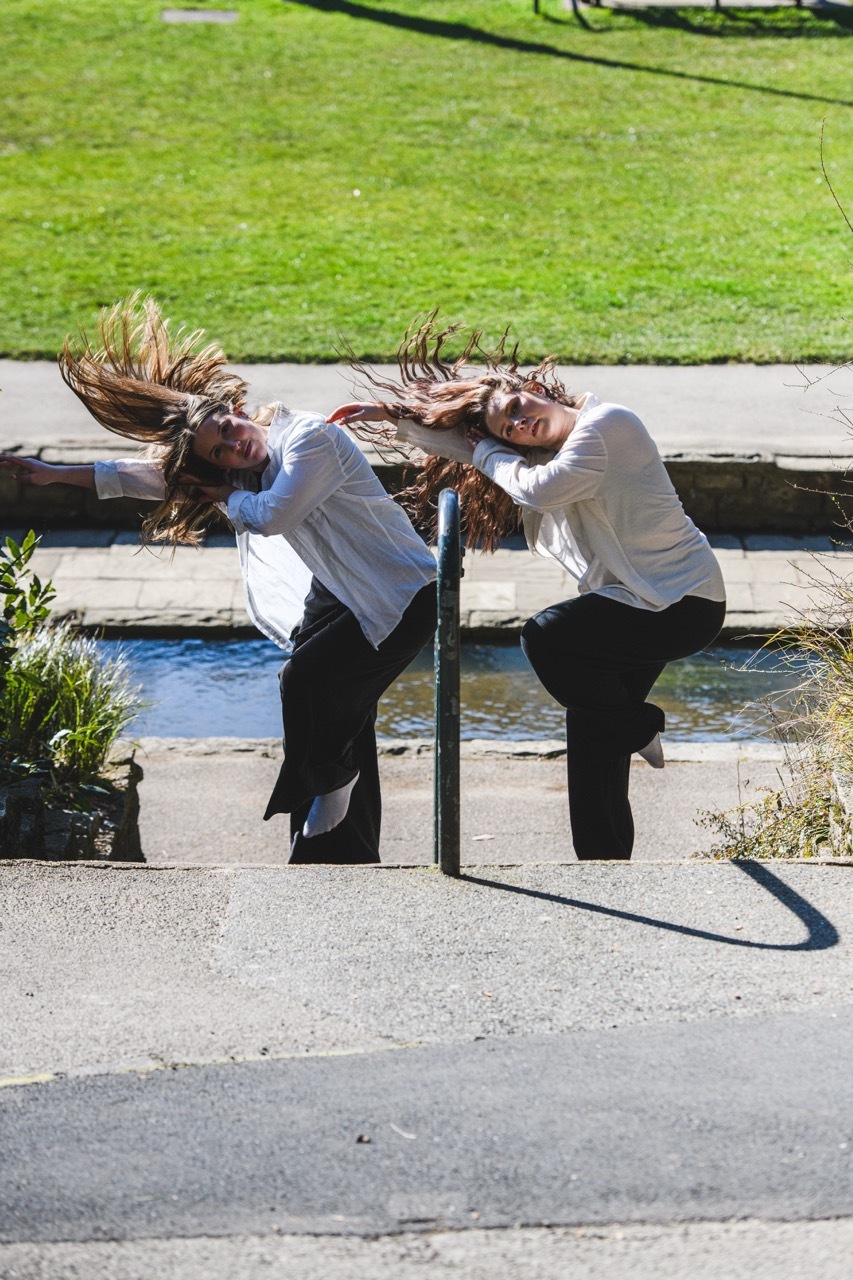 Two women with long hair in motion, wearing white tops and dark trousers, positioned on either side of a vertical pole on concrete pavement.