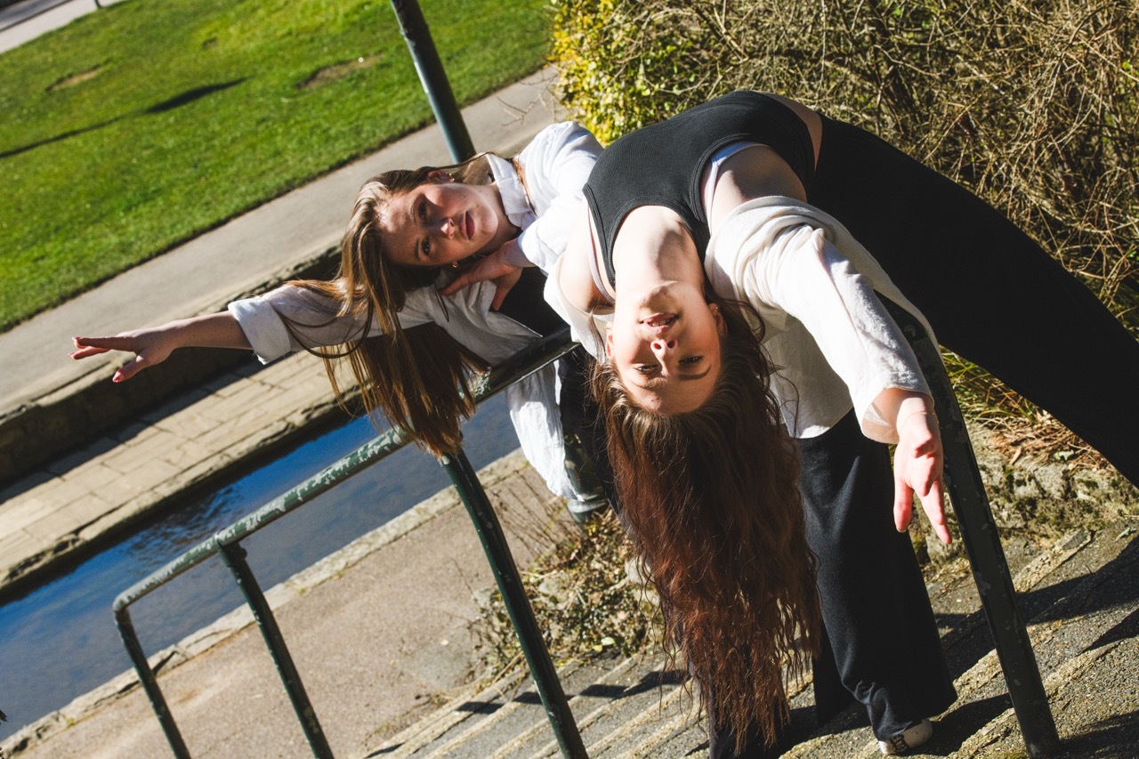 Two women hanging upside down on playground equipment, smiling at camera with green grass and concrete visible in background.
