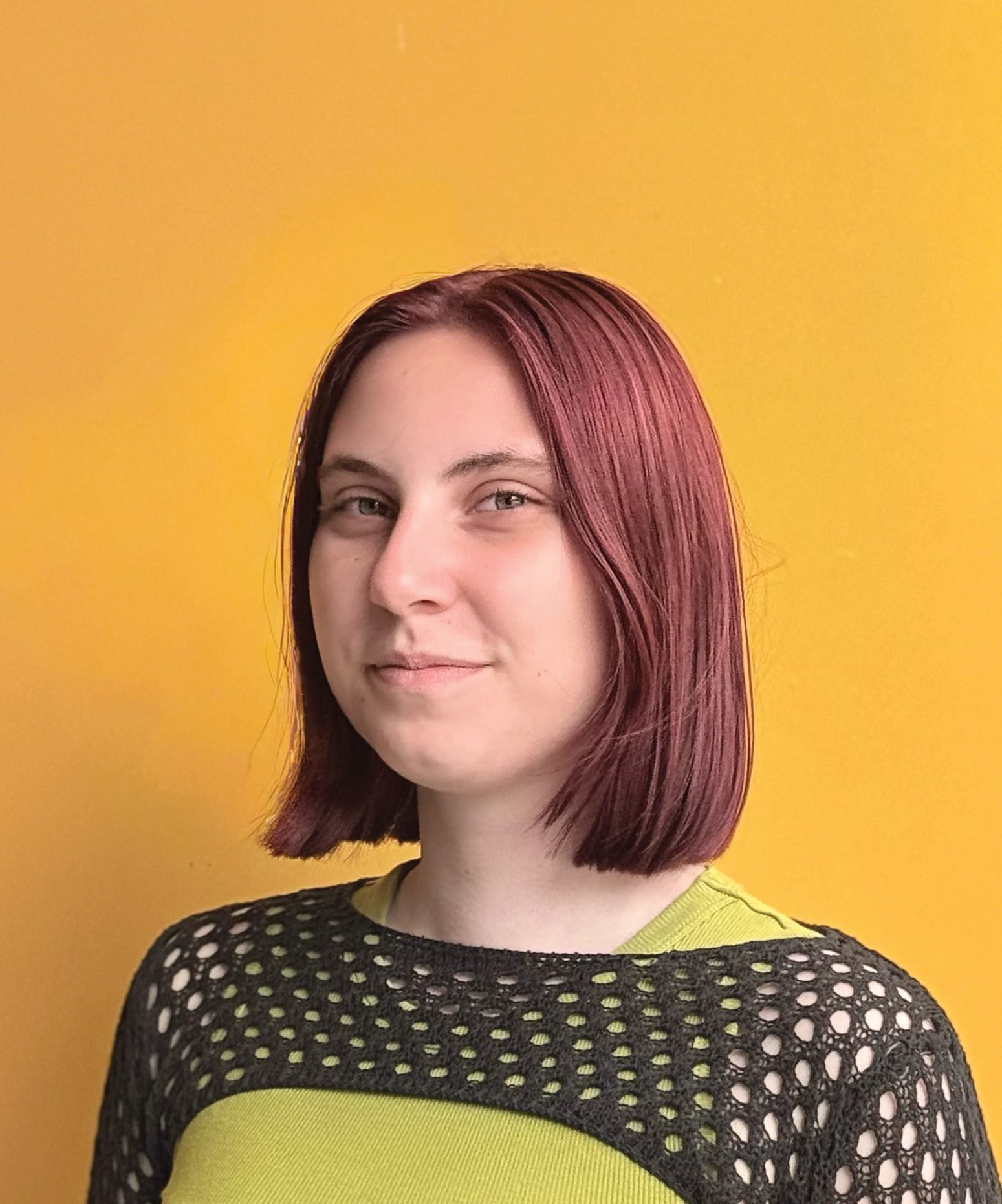 Woman with shoulder-length reddish-brown bob haircut wearing green top with black mesh overlay against bright orange background.