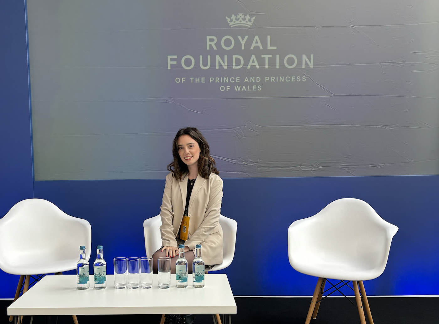 Headshot of A. Shipton sitting by a coffee table with the Royal Foundation logo on the wall behind them.