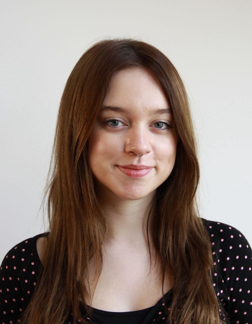 Young woman with long brown hair wearing black polka dot top, smiling slightly against white wall background.