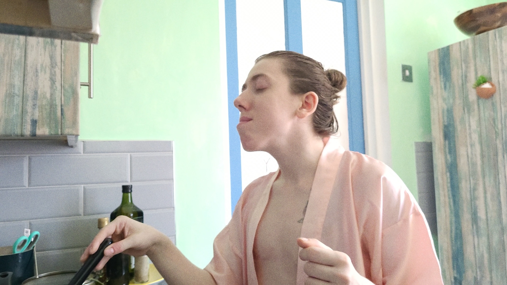 A woman in a light pink robe cooks at a stove in a bright kitchen with green walls and blue trim.