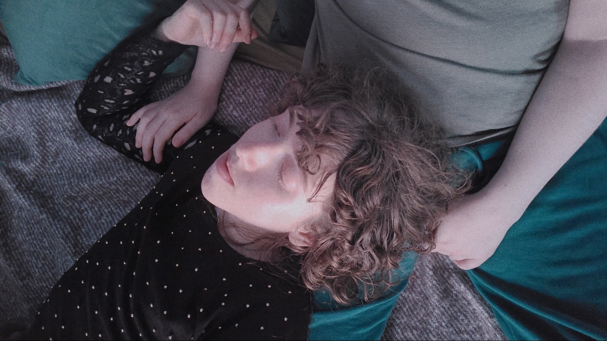 A person with curly hair lies on a grey sofa, resting their head on another person's lap, with teal cushions and a patterned sleeve visible nearby.