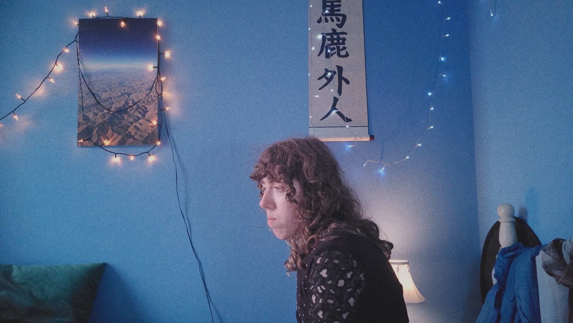 A young person with curly hair sits in profile in a blue-lit room, with string lights, a landscape poster and a vertical banner on the wall behind them.