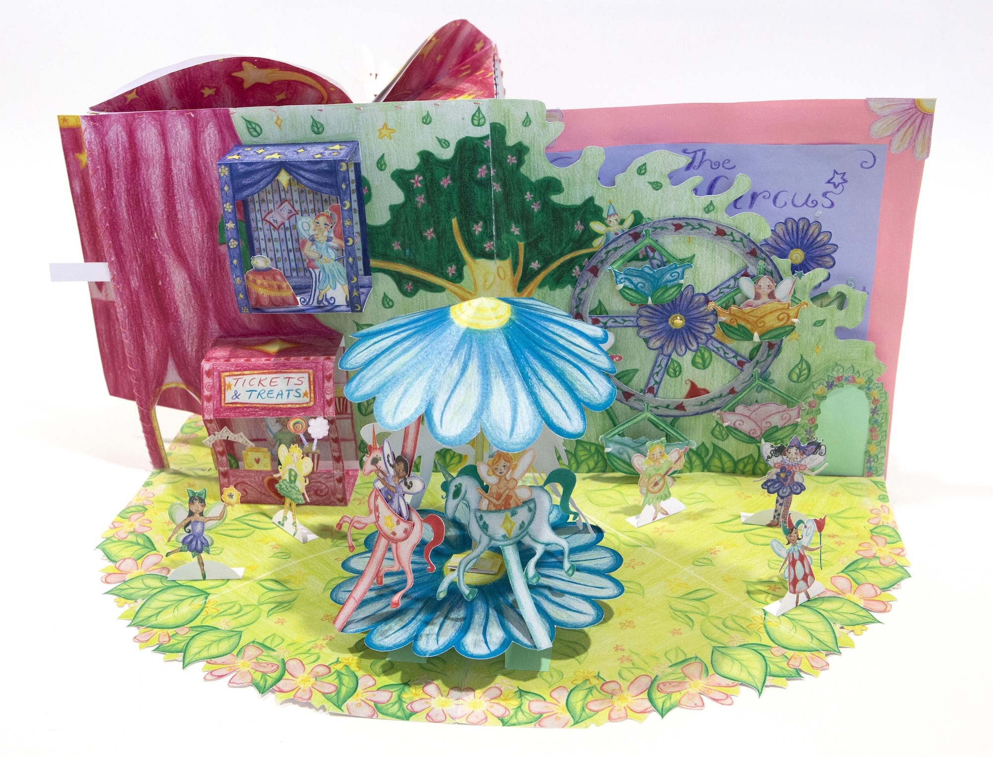 Pop-up book showing fairy garden scene with blue flower centre piece, green landscape, pink building backdrop, and small figures.