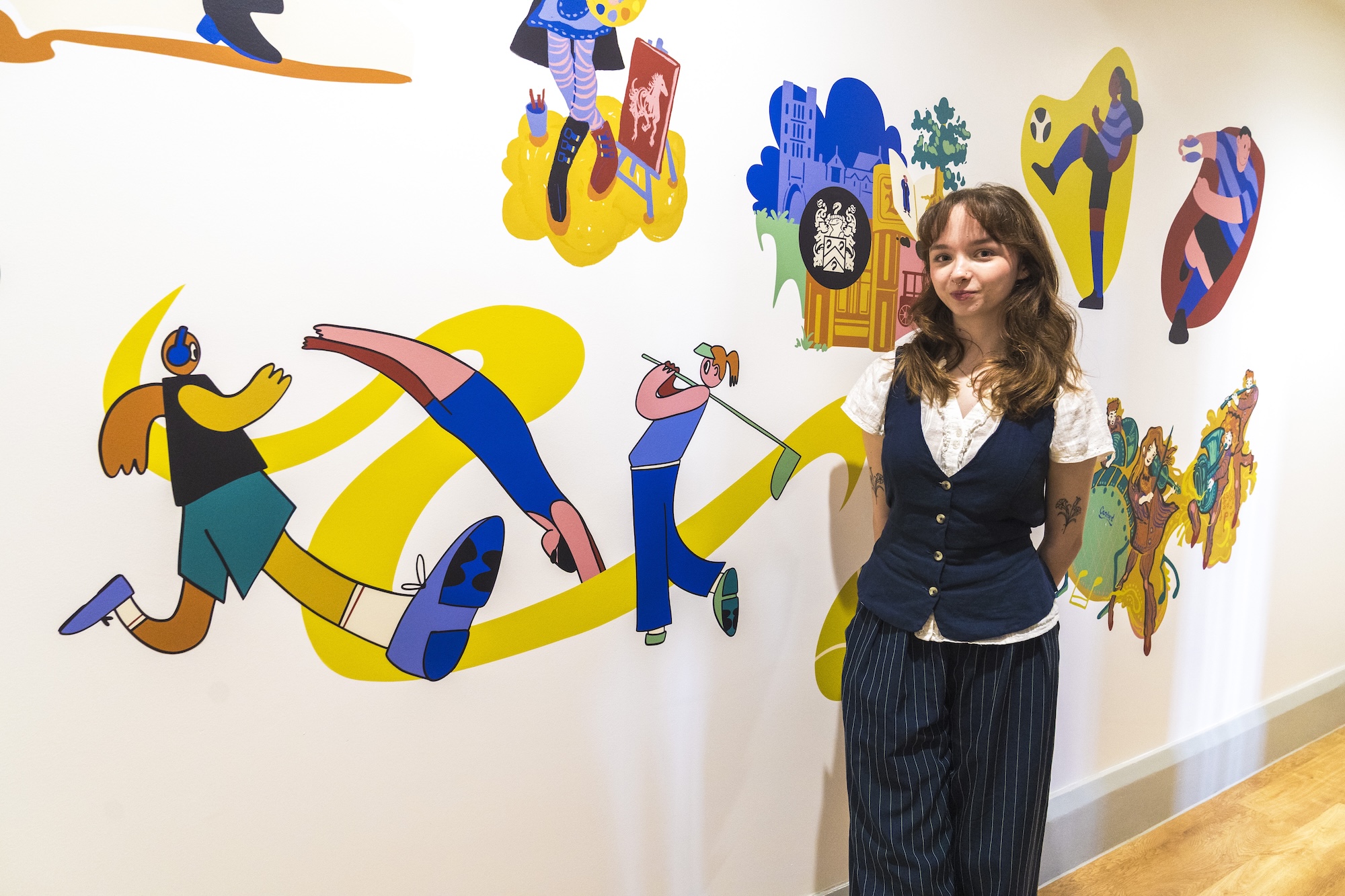 BA (Hons) Illustration student A. Ryalls stands next to several colourful vinyl illustrations on a white wall and poses for a photo.