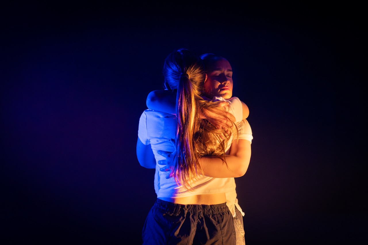 Two young women are embracing each other against a dark background. One is wearing a white top and the other a black outfit, while soft lighting in shades of blue and purple highlights their figures.