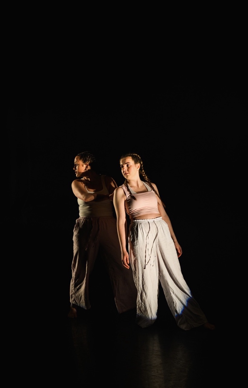 Two dancers are positioned in a dark space, with one facing away and the other slightly turned towards the camera. Both are wearing light-coloured tops and loose, flowing trousers, illuminated by a soft spotlight that highlights their forms.