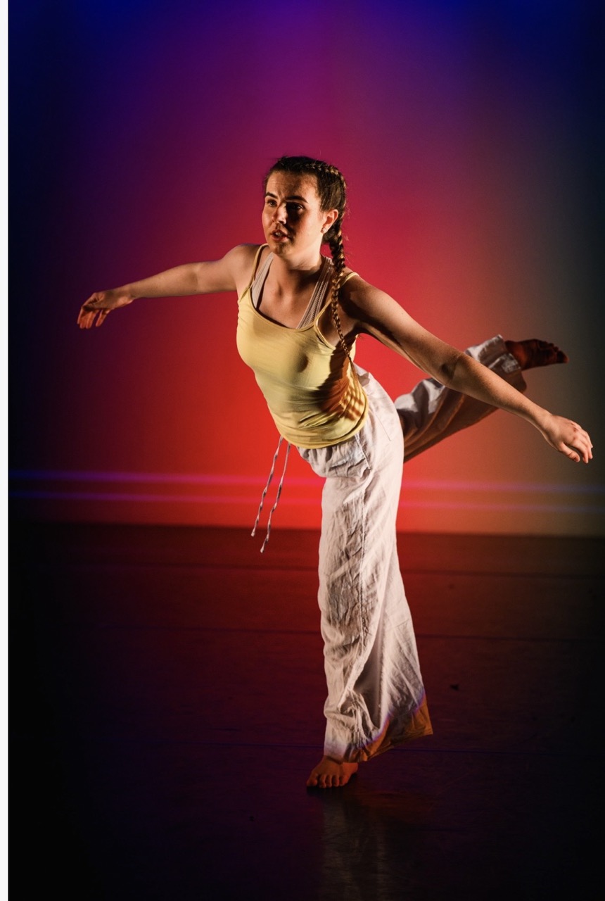 A young woman balances on one foot in a dance pose, wearing a yellow top and flowing white trousers. The background features a gradient of red and purple lighting, creating a vibrant atmosphere.