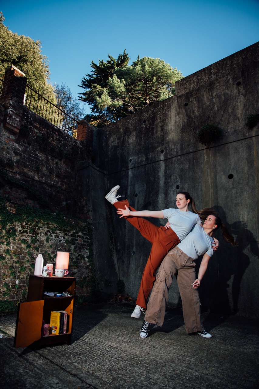 Two women engaged in a playful dance pose in an outdoor setting. One woman is lifting the leg of the other who is standing beside her, with a small table featuring books and a lamp in the foreground. A wall and trees are visible in the background.