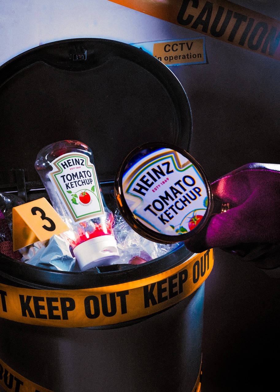 A modern trash bin with a yellow caution tape around it. Inside the bin, there are two bottles of Heinz tomato ketchup, partially submerged in ice. A magnifying glass is held above the bottle, with a label featuring the product name visible.