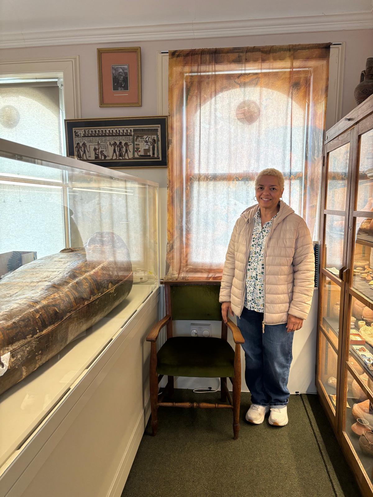 Woman in beige jacket stands in museum room with glass display cases containing artefacts and mummy, framed artwork on walls.