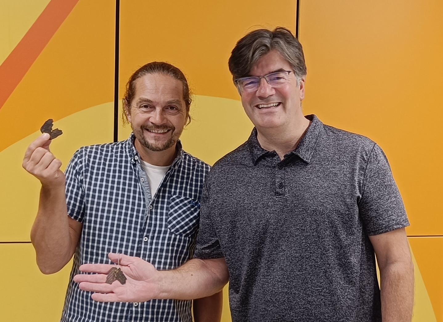 Two men stand in front of an orange wall with a curved yellow stripe, one holding small dark objects in his hand and the other standing beside him in glasses.