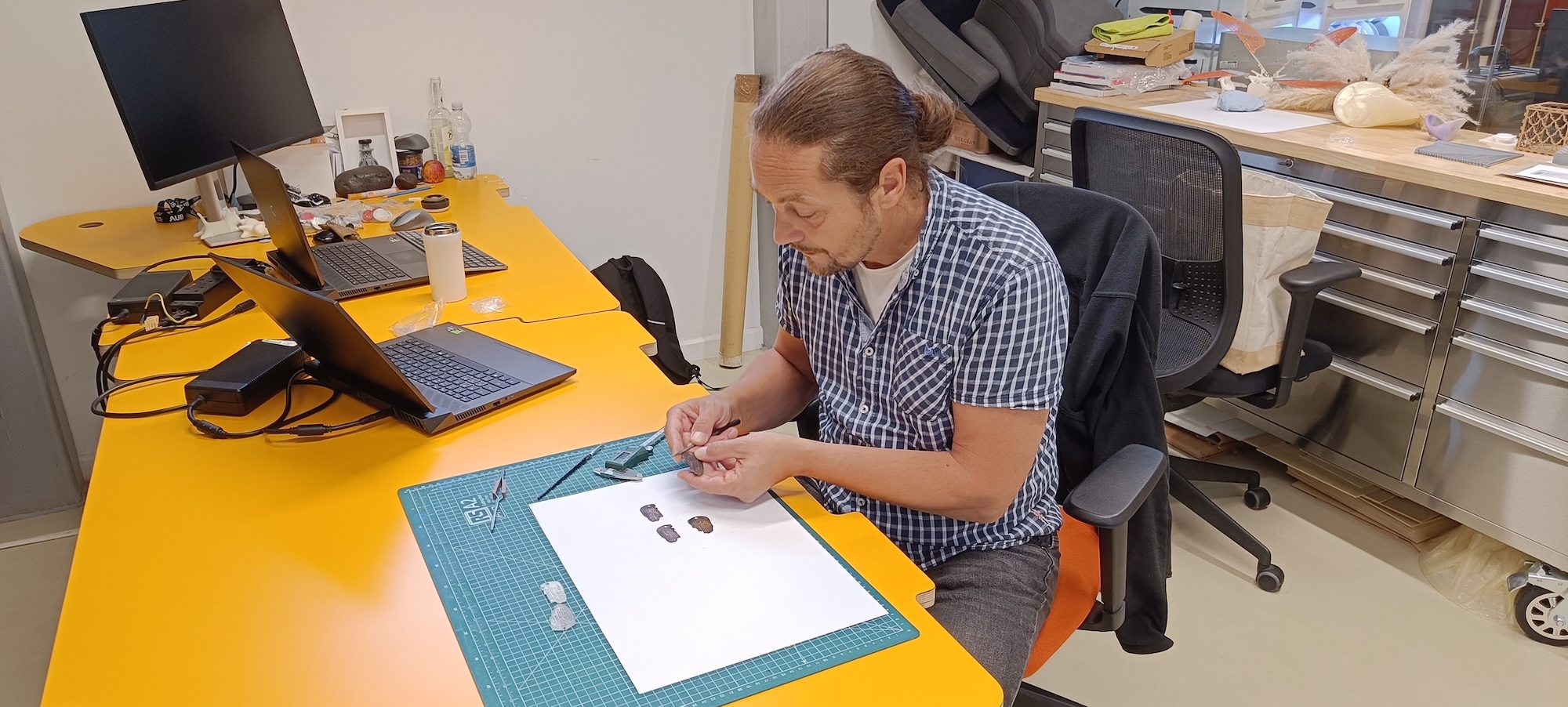 A man sits at an orange desk sorting small coin-like objects on a cutting mat, with a laptop and monitor beside him in a bright office.