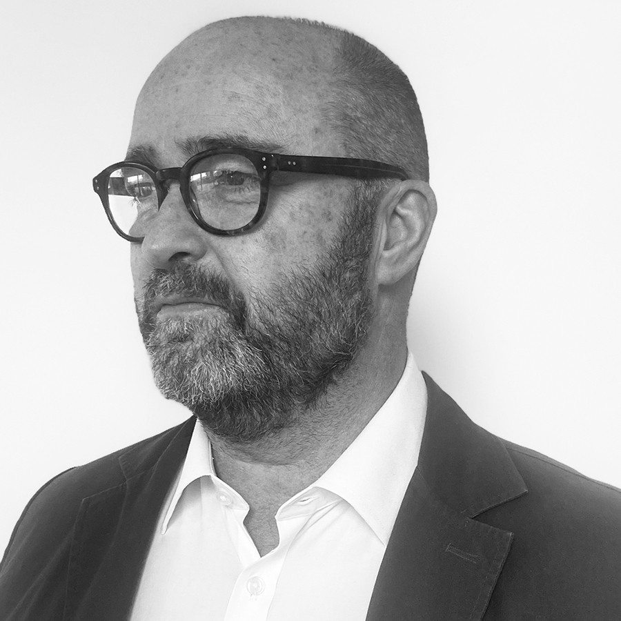 Middle-aged bald man with thick-rimmed glasses and full beard wearing dark suit jacket over white shirt, photographed in black and white.