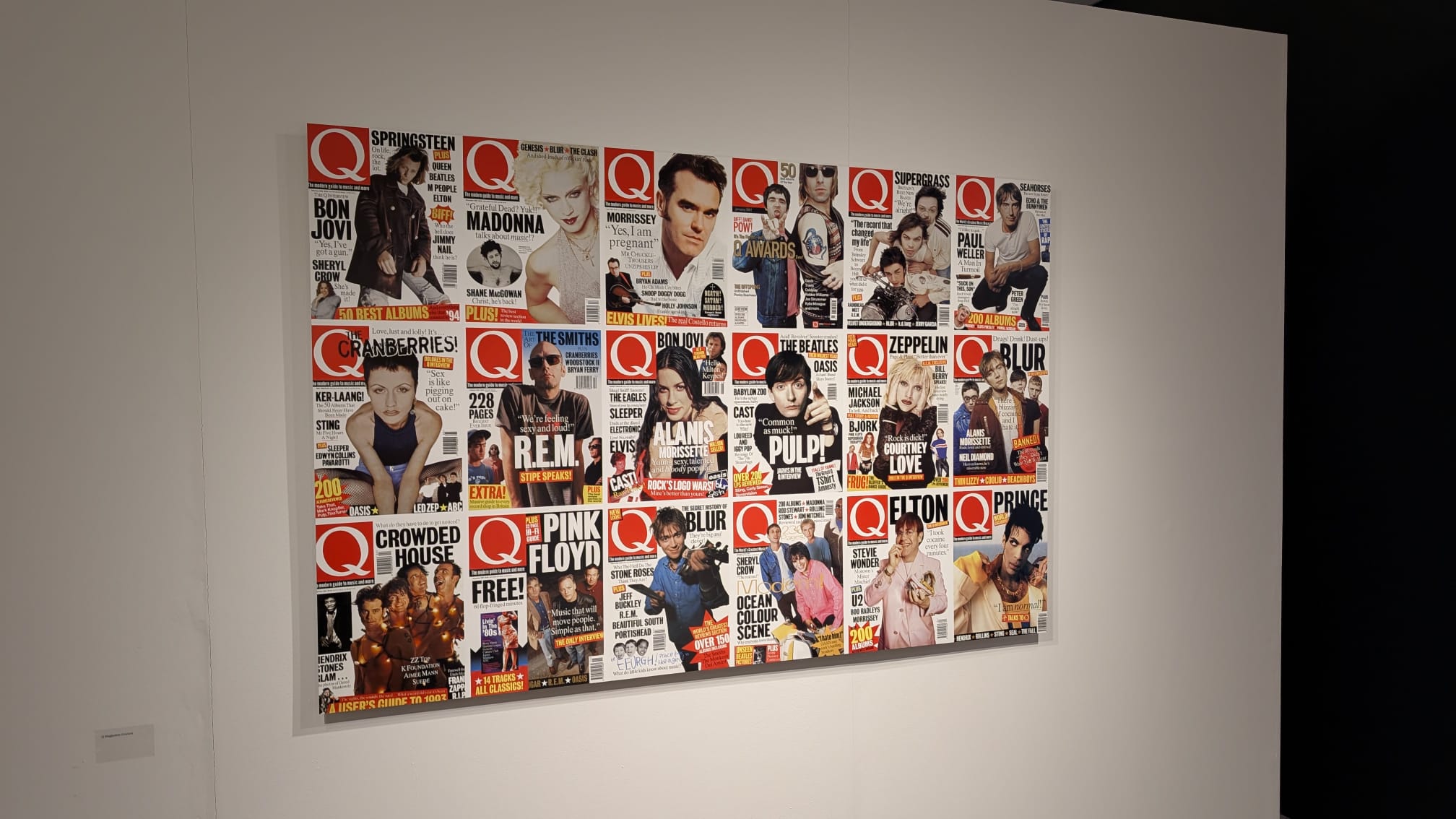 A framed collage of Q magazine covers featuring musicians and bands, mounted on a white gallery wall with a small label beneath it.